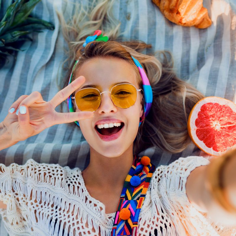 Excited girl making selfie on beach towel. Boho accessories. Yellow sunglasses.Top view.