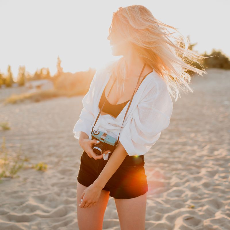 Stylish shapely girl with retro camera posing on sunny evening beach. Tropical mood.